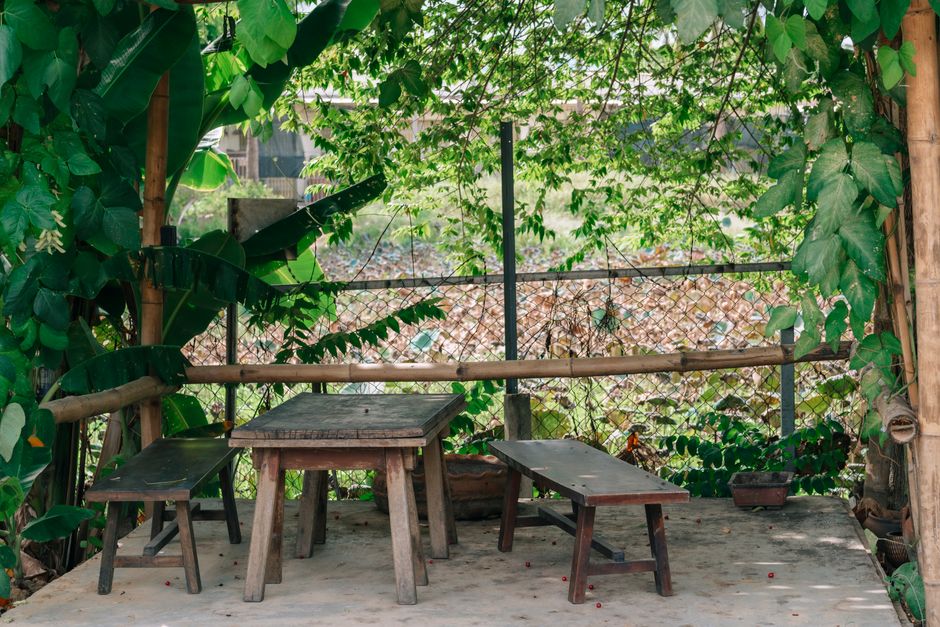 A small, rustic outdoor garden area featuring a wooden table and two matching benches, with additional small stools placed nearby. The space is partially enclosed by a wooden frame and a wire mesh fen